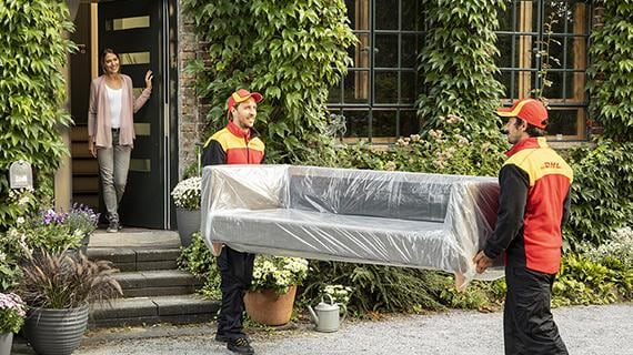 Zwei Männer in DHL-Kleidung tragen ein Sofa in das Haus einer Frau