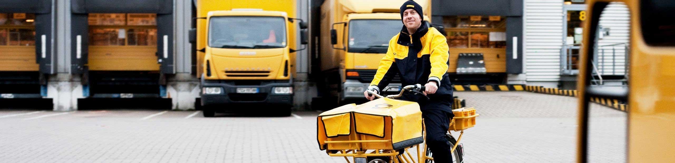 Ein lächelnder Postbote der Deutschen Post sitzt auf einem Zustellfahrrad, im Hintergrund sind zwei LKWs an Laderampen zu sehen.