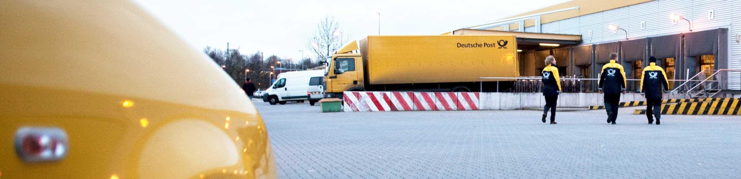 Drei Mitarbeiter der Deutschen Post laufen auf eine Zustellbasis zu, vor der ein LKW der Deutschen Post an einer Laderampe steht.