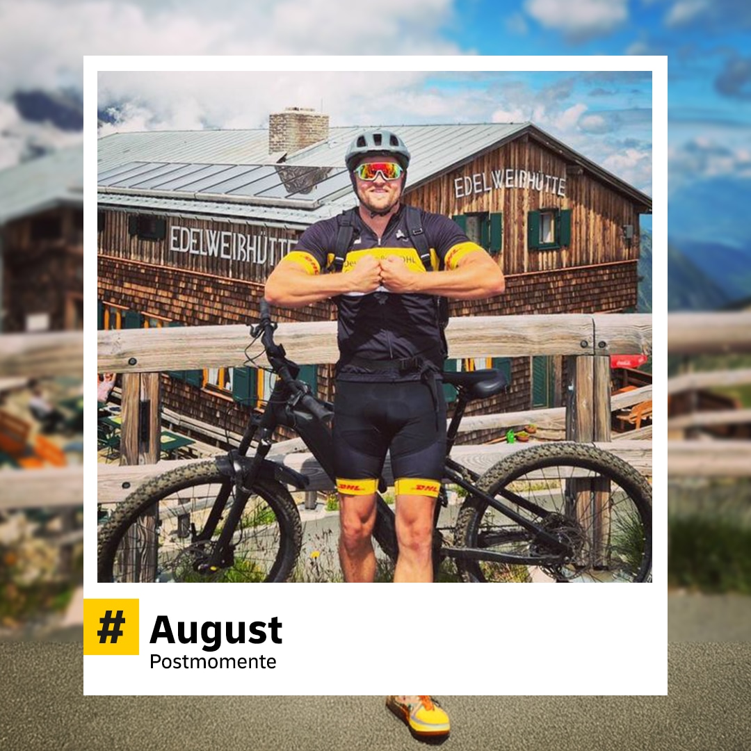 Sportlicher Postmitarbeiter mit Helm steht vor einem Fahrrad in den Bergen und hält ein Paket. Im Hintergrund eine Almhütte. Hashtag: „#August – Postmomente“.