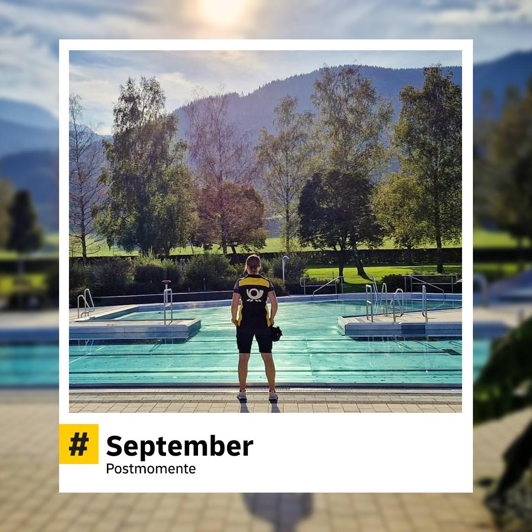 Postmitarbeiter:in steht auf einem Bootssteg an einem ruhigen See mit Bergen im Hintergrund. Strahlt Sommer-Idylle aus. Hashtag: „#September – Postmomente“.