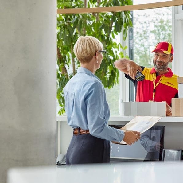 Zusteller in DHL-Uniform überreicht einer Kundin in moderner Filiale ein Paket am Schalter. Die Kundin trägt eine hellblaue Bluse und steht an einem Empfang.