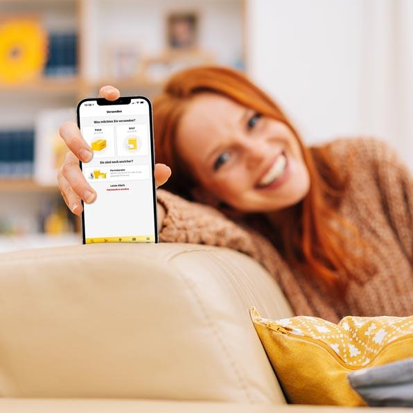 Zwei Frauen sitzen entspannt auf dem Sofa. Eine hält ein Smartphone mit der geöffneten Post & DHL App in die Kamera.