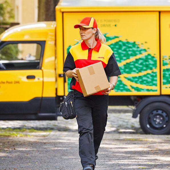 Zustellerin der Deutschen Post in gelber Uniform mit roter Mütze trägt ein Paket vor einem GoGreen Plus-Lieferwagen mit grünem Blätterdesign.