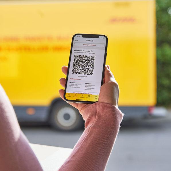 Hand hält ein Smartphone mit digitaler Paketmarke vor einem gelben DHL-Transporter. Auf dem Display ist ein QR-Code sichtbar.