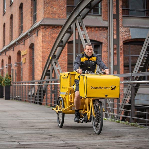 Zusteller der Deutschen Post fährt mit einem gelben Lastenfahrrad über eine Brücke in einem urbanen Industriegebiet. Er lächelt in die Kamera.