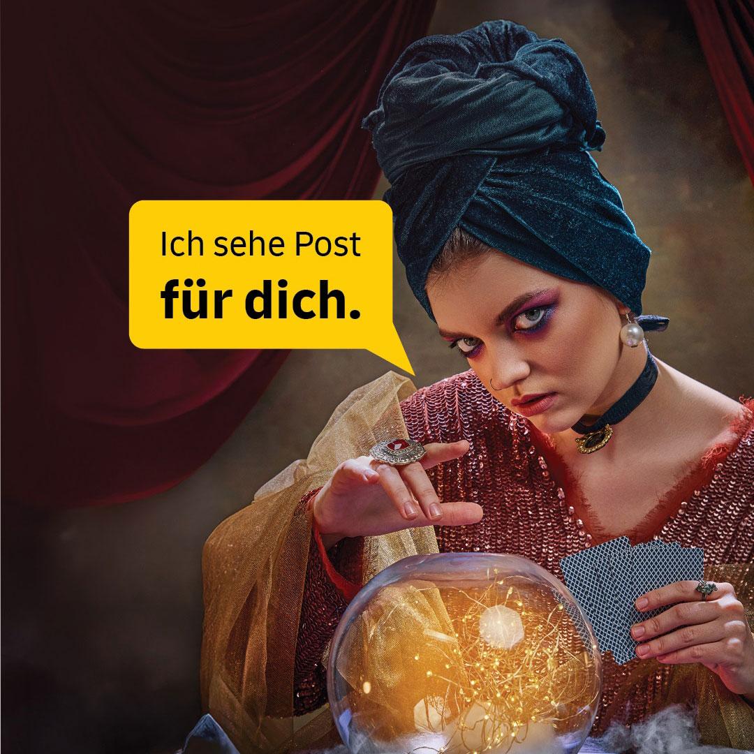 Eine Wahrsagerin mit Turban und aufwendigem Make-up schaut geheimnisvoll in eine leuchtende Glaskugel, während sie Karten hält. Gelbe Sprechblase im Bild: „Ich sehe Post für dich.“